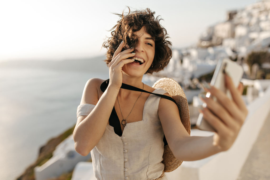 Brunette curly woman in beige dress and straw hat smiles sincerely and takes selfie outside in old